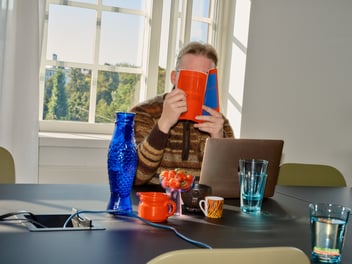 alt text: man at a desk with glass of water, in front of a laptop holding a book in front of his face. 
