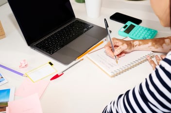 Person writing notes in front of a laptop