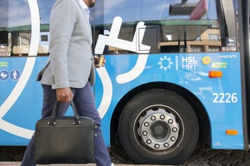 A passenger carrying a case walking in front of a blue HSL bus. HSL logo and an image of a large electric socket is pictured on the side of the bus.